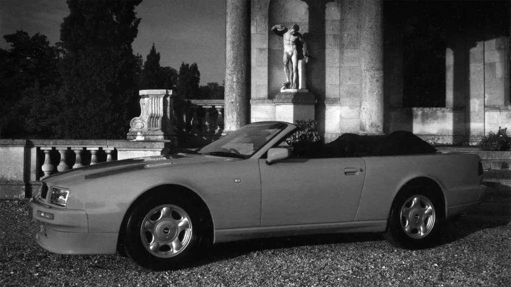 Aston-Martin-Virage-Volante-outside-stately-home-hood-down-large-stone-sculpture-with columns-on-house